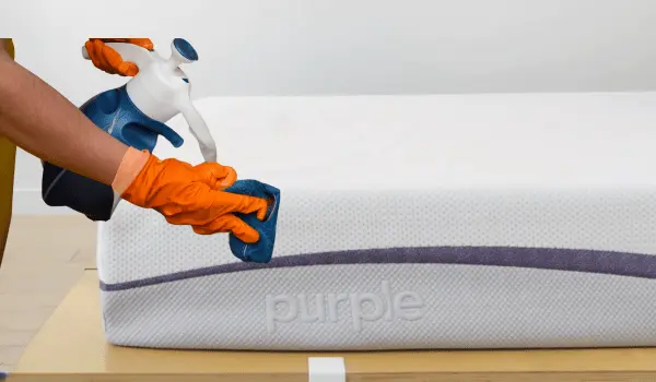 A person wearing gloves spraying and wiping a Purple Mattress with a cleaning solution.