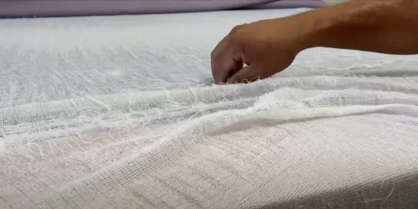 Close-up of a hand examining the inner fabric layers of a mattress to check for the presence of fiberglass fibers.