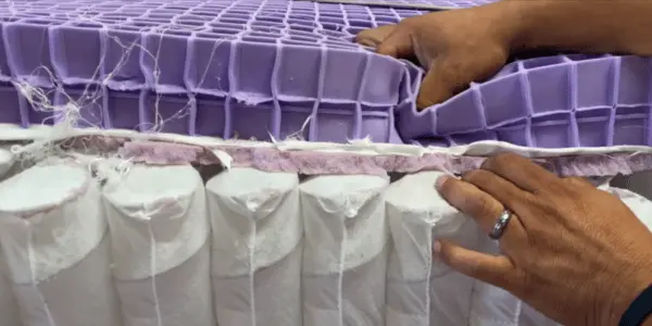 Hands revealing the different construction layers of a Purple mattress, including the hyper-elastic polymer grid and foam layers.