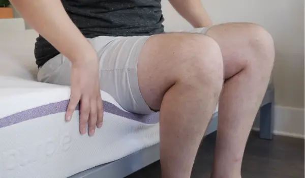 A person sitting on the edge of the Purple Mattress demonstrates its edge support and checks if the edges feel weaker or more prone to collapsing.